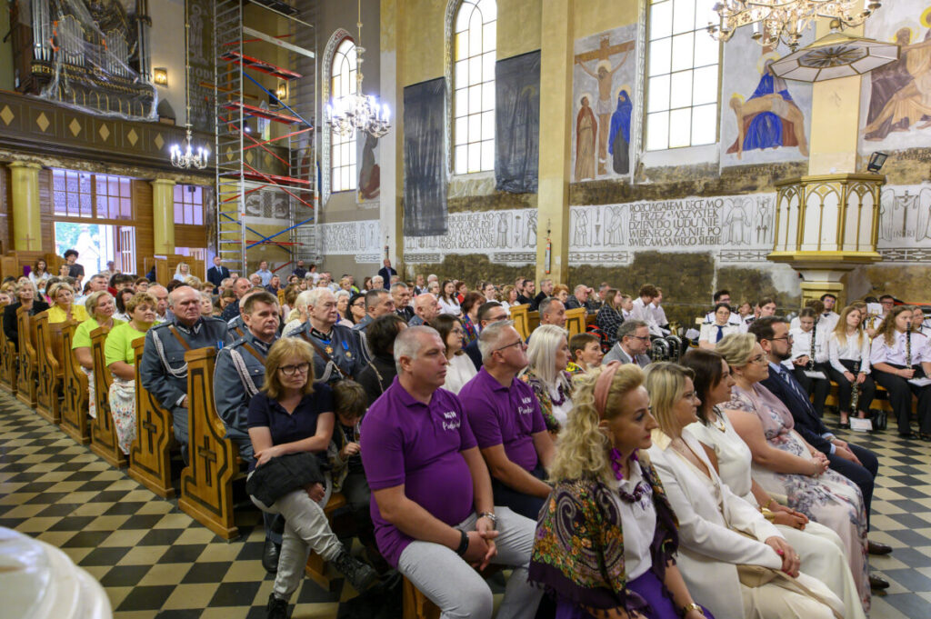 Wnętrze kościoła, ludzie siedzą w ławkach. Ściany zdobią freski, a światło pada przez duże okna. Tło jasne, sufit z żyrandolem.