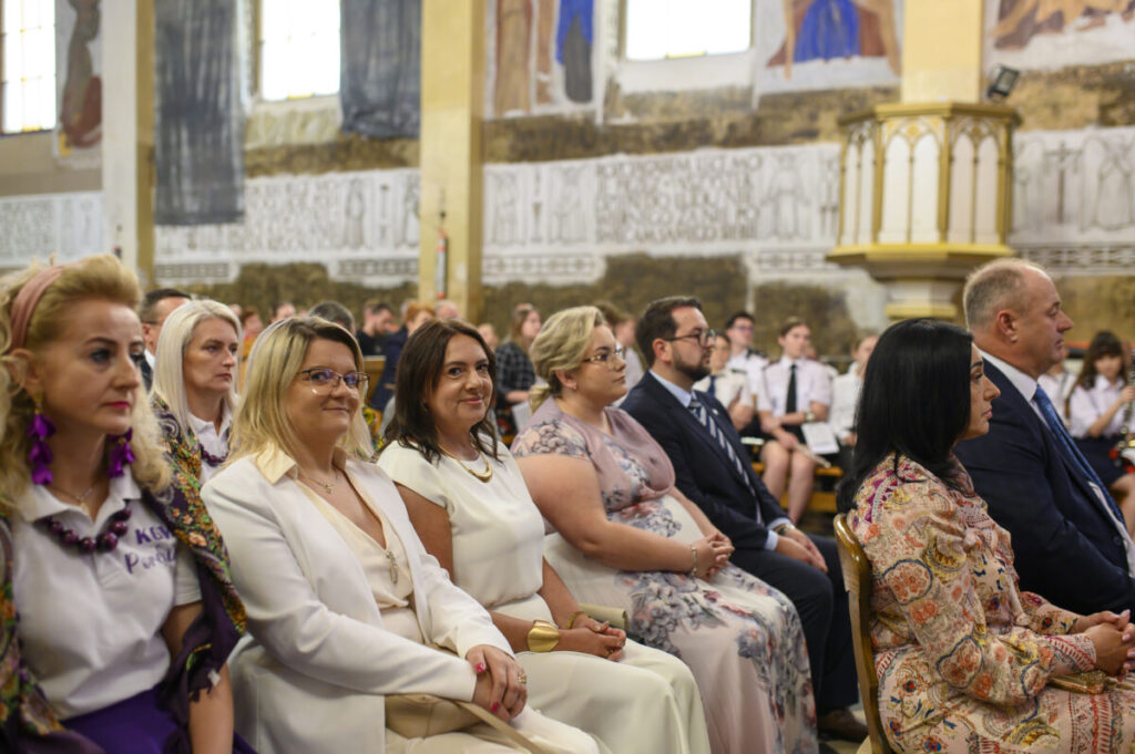 Sala z uczestnikami w eleganckich strojach siedzącymi w rzędach. Jasne wnętrze z ozdobnymi detalami na ścianach. Grupa skupiona na wydarzeniu.