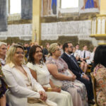 Sala z uczestnikami w eleganckich strojach siedzącymi w rzędach. Jasne wnętrze z ozdobnymi detalami na ścianach. Grupa skupiona na wydarzeniu.