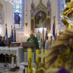 Wnętrze kościoła z mężczyzną w zielonym ornacie przy ambonie. W tle kolorowe witraże i obrazy. Z boku stoją flagi i strażacy w mundurach.