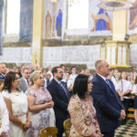 Grupa ludzi w eleganckich strojach stoi w kościele. Ściany zdobią freski w jasnych kolorach. Na suficie wisi żyrandol.