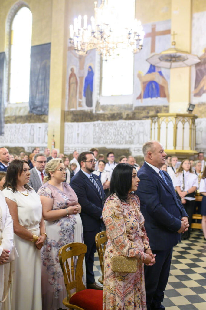 Grupa ludzi w eleganckich strojach stoi w kościele. Ściany zdobią freski w jasnych kolorach. Na suficie wisi żyrandol.