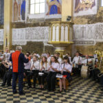 Orkiestra dęta w kościele, jasne wnętrze z freskami. Muzycy w białych koszulach grają, dyrygent w ciemnym stroju. Czarno-biała posadzka.