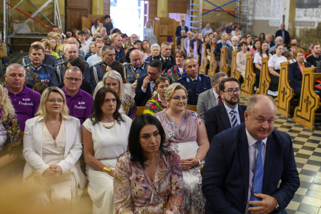 Zdjęcie przedstawia grupę ludzi w jasnym wnętrzu kościoła. Ludzie siedzą na drewnianych ławkach, ubrani w eleganckie stroje. W tle widać dekoracyjne detale ścian i podłogi w szachownicę.