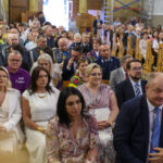 Zdjęcie przedstawia grupę ludzi w jasnym wnętrzu kościoła. Ludzie siedzą na drewnianych ławkach, ubrani w eleganckie stroje. W tle widać dekoracyjne detale ścian i podłogi w szachownicę.