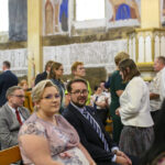 Grupa ludzi siedzi w kościele, ubrani elegancko. Ściany zdobią freski w ciepłych kolorach. Wokół panuje atmosfera skupienia.