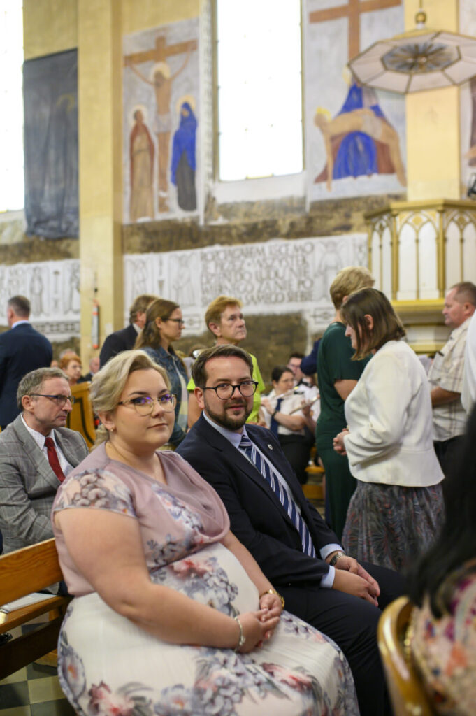 Grupa ludzi siedzi w kościele, ubrani elegancko. Ściany zdobią freski w ciepłych kolorach. Wokół panuje atmosfera skupienia.