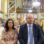 Ludzie siedzą w kościele z rusztowaniami w tle. Mężczyzna w garniturze i kobieta w kolorowej sukience. Jasne oświetlenie wnętrza.