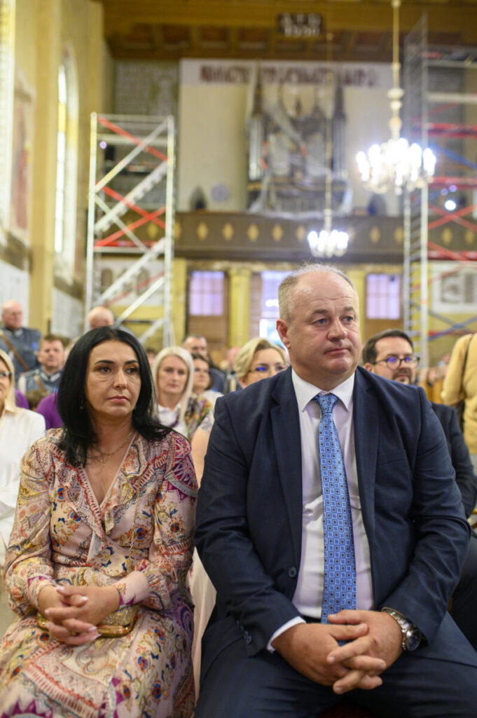 Ludzie siedzą w kościele z rusztowaniami w tle. Mężczyzna w garniturze i kobieta w kolorowej sukience. Jasne oświetlenie wnętrza.