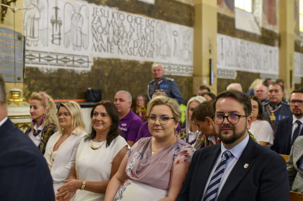 Grupa elegancko ubranych ludzi siedzi we wnętrzu kościoła z inskrypcjami na ścianie. Dominują kolory białe i złote. Tło zawiera napisy i elementy religijne.