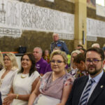 Grupa elegancko ubranych ludzi siedzi we wnętrzu kościoła z inskrypcjami na ścianie. Dominują kolory białe i złote. Tło zawiera napisy i elementy religijne.