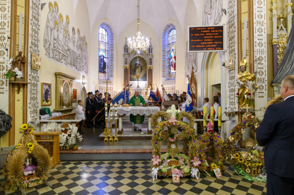 Kościół z ołtarzem, dekoracjami i sztandarami. W tle witraże i freski na ścianach. Przeważają barwy złote i kremowe.