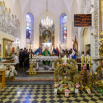 Kościół z ołtarzem, dekoracjami i sztandarami. W tle witraże i freski na ścianach. Przeważają barwy złote i kremowe.