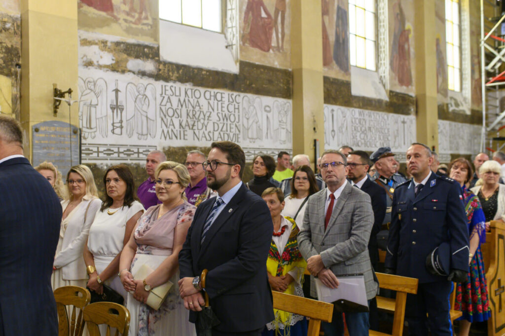 Grupa ludzi stoi w kościele z freskami na ścianach. Tło jest jasne z białymi i złotymi odcieniami. Wszyscy są odświętnie ubrani.