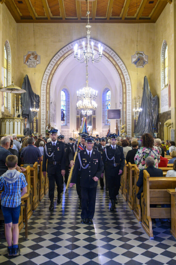 Grupa mężczyzn w mundurach maszeruje w kościele z witrażami. Podłoga w szachownicę, złote detale architektury. Eleganckie żyrandole świecą nad tłumem.