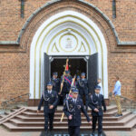 Grupa strażaków w mundurach wychodzi z ceglanego kościoła. Na fasadzie widoczny biały krzyż i obraz Matki Boskiej. Tło stanowi czerwono-brązowa cegła z białymi akcentami.