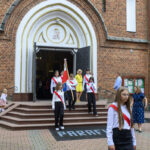 Przed kościołem z cegły wychodzi grupa młodzieży z flagą. Mają białe koszule i czerwone szarfy. Na tle drzwi widoczna jest postać Matki Boskiej.