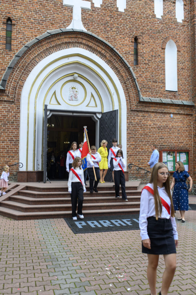 Przed kościołem z cegły wychodzi grupa młodzieży z flagą. Mają białe koszule i czerwone szarfy. Na tle drzwi widoczna jest postać Matki Boskiej.
