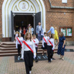 Na zdjęciu dzieci w białych koszulach i z czerwonymi szarfami opuszczają kościół z ceglanym frontem. Na pierwszym planie widoczne są schody, a w tle otwarte drzwi kościoła. Dorośli idą za nimi, po obu stronach t