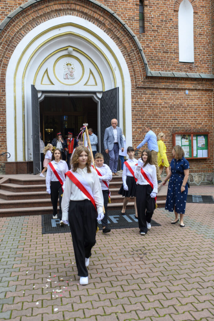 Na zdjęciu dzieci w białych koszulach i z czerwonymi szarfami opuszczają kościół z ceglanym frontem. Na pierwszym planie widoczne są schody, a w tle otwarte drzwi kościoła. Dorośli idą za nimi, po obu stronach t