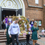 Grupa ludzi w uroczystym ubiorze wychodzi z kościoła z czerwonej cegły. Niosą dekoracyjny wieniec z kwiatami. Tło zdobią drzwi kościoła i tekst IHS.