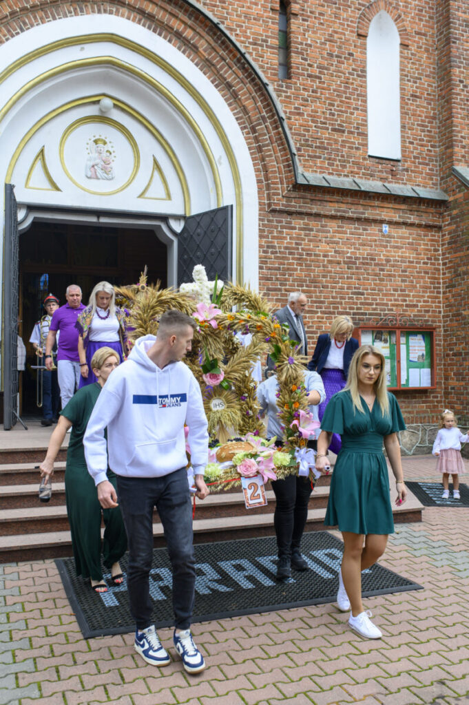 Grupa ludzi w uroczystym ubiorze wychodzi z kościoła z czerwonej cegły. Niosą dekoracyjny wieniec z kwiatami. Tło zdobią drzwi kościoła i tekst IHS.