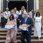 Grupa elegancko ubranych ludzi wychodzi z kościoła, trzymając chleb na białych serwetkach. Są na schodach z czerwonego kamienia, tle widoczny biały portal z wizerunkiem Matki Boskiej.