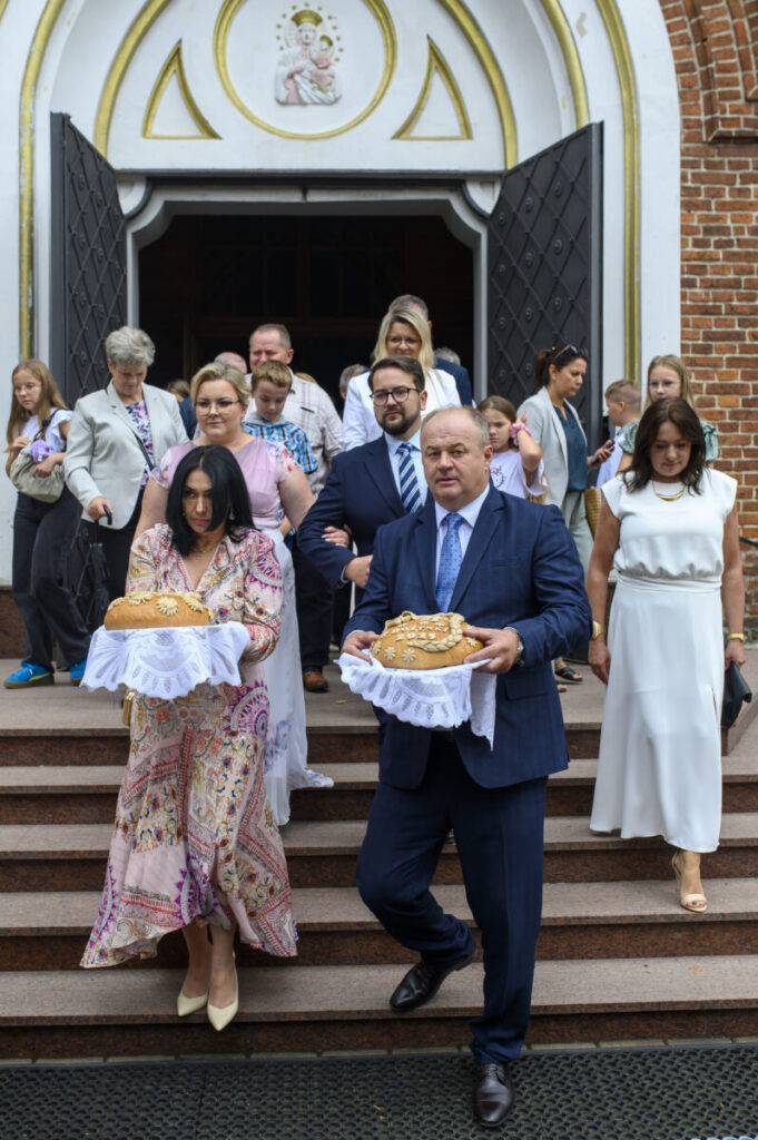 Grupa elegancko ubranych ludzi wychodzi z kościoła, trzymając chleb na białych serwetkach. Są na schodach z czerwonego kamienia, tle widoczny biały portal z wizerunkiem Matki Boskiej.
