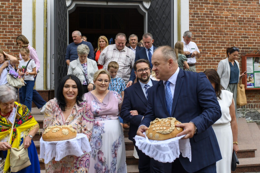 Grupa ludzi wychodzi z kościoła, trzymając tradycyjne chleby. Budynek z cegły w tle. Kobiety w kolorowych strojach.