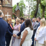 Grupa ludzi elegancko ubranych stoi przed ceglanym kościołem otoczonym drzewami. Dwie kobiety w centrum uśmiechają się, trzymając się za ręce. Tło jest zielone i widać inne osoby oraz drzwi kościoła.