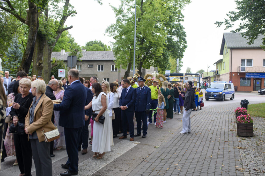Grupa ludzi stoi w kolejce na ulicy, otoczona drzewami. W tle niebieski policyjny pojazd na tle budynków. Przeważają zielone i szare kolory.