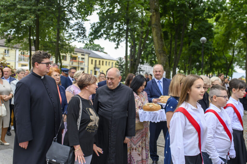 Na zdjęciu jest zbieg, grupa osób, m.in. duchowni i dzieci w strojach galowych, na tle zielonego parku. Dzieci niosą chleb na ozdobnych tacach. W tle widoczne są drzewa i budynki.