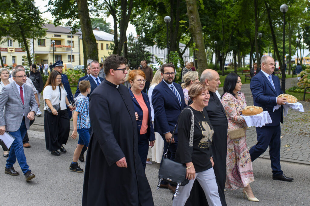 Grupa osób idzie przez park z bujną zielenią na tle budynków. Dwóch mężczyzn niesie chleby na białych serwetach. Wszyscy ubrani elegancko, atmosfera jest uroczysta.