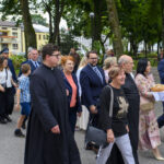 Grupa osób idzie przez park z bujną zielenią na tle budynków. Dwóch mężczyzn niesie chleby na białych serwetach. Wszyscy ubrani elegancko, atmosfera jest uroczysta.