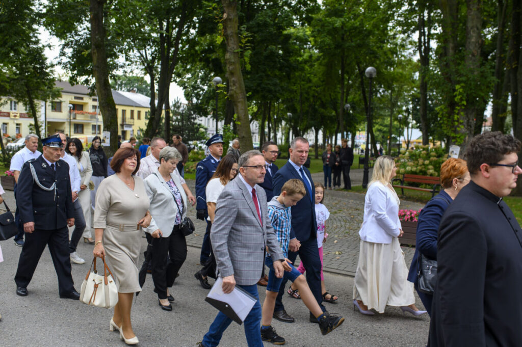 Grupa ludzi spaceruje alejką w parku, ubrani formalnie. W tle drzewa i budynki. Atmosfera jest spokojna i elegancka.
