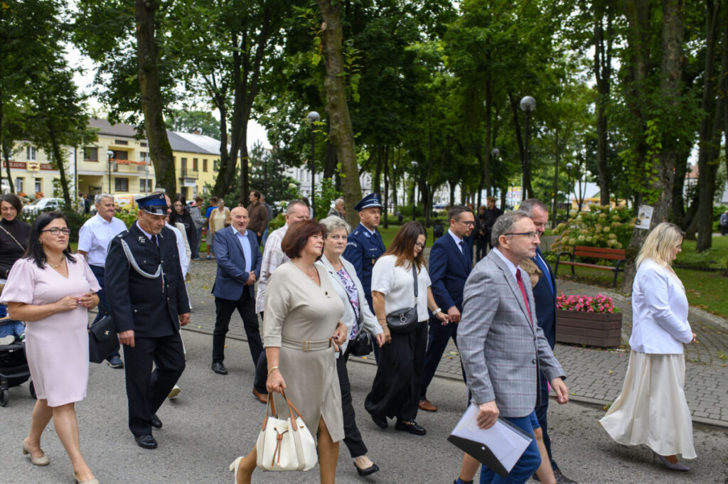 Grupa ludzi, w tym strażak, spaceruje po parku. W tle widać drzewa i budynki. Dominuje zieleń i stonowane kolory ubrań.