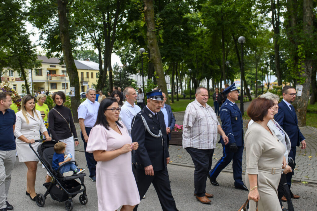 Grupa ludzi, w tym policjant, maszeruje alejką w parku. Otaczają ich wysokie drzewa, a w tle widoczne są budynki. Kolory są żywe, atmosfera spokojna.
