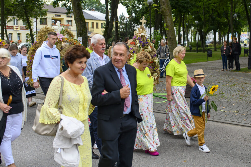 Na zdjęciu widać grupę ludzi biorących udział w procesji z wieńcami i słonecznikami. Dominują kolory zielony, żółty i beżowy. Tło stanowi park z drzewami i brukowaną alejką.