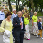 Na zdjęciu widać grupę ludzi biorących udział w procesji z wieńcami i słonecznikami. Dominują kolory zielony, żółty i beżowy. Tło stanowi park z drzewami i brukowaną alejką.