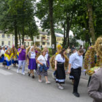Ludzie idą w procesji przez miasto, niosąc kolorowe wieńce z kwiatów. Ubrani są w tradycyjne stroje, dominują fioletowe i żółte barwy. W tle widać budynki i drzewa.