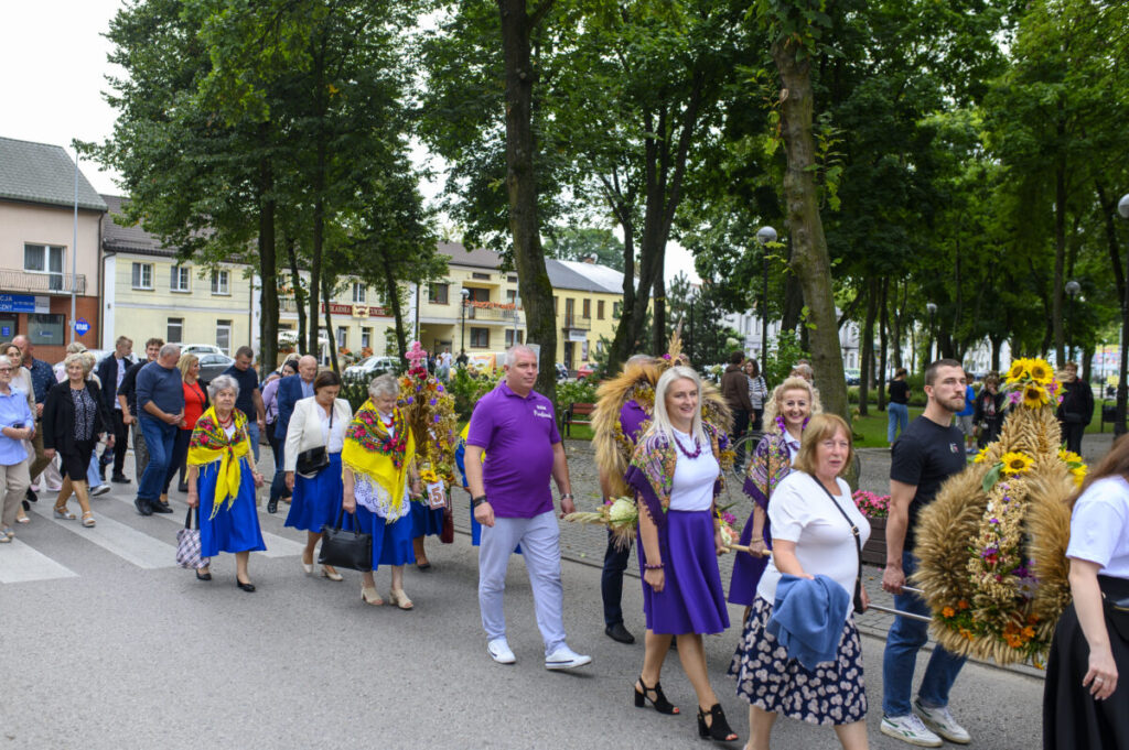 Grupa ludzi idąca w procesji w parku, ubrana w kolorowe stroje ludowe z kwiatowymi dekoracjami. Tło to drzewa i budynki w jasnych barwach. Wydarzenie ma charakter tradycyjny.