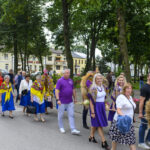 Grupa ludzi idąca w procesji w parku, ubrana w kolorowe stroje ludowe z kwiatowymi dekoracjami. Tło to drzewa i budynki w jasnych barwach. Wydarzenie ma charakter tradycyjny.