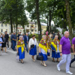 Grupa osób w kolorowych strojach idzie w procesji po ulicy, z domami i drzewami w tle. Uczestnicy niosą ozdobne wieńce, a kobiety mają żółte chusty. Niebo jest zachmurzone.