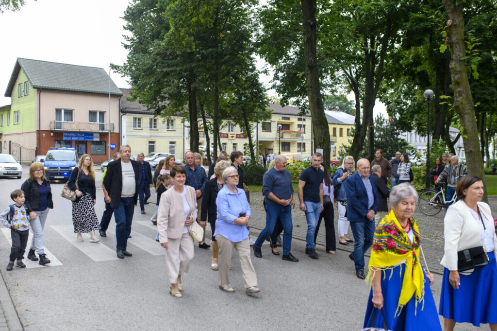 Grupa ludzi spaceruje w parku miejskim pełnym zieleni. W tle widać budynki w jasnych kolorach. Uczestnicy ubrani różnorodnie, część w jaskrawych strojach.