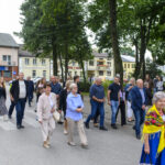 Grupa ludzi spaceruje w parku miejskim pełnym zieleni. W tle widać budynki w jasnych kolorach. Uczestnicy ubrani różnorodnie, część w jaskrawych strojach.