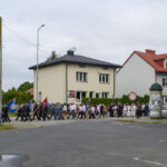 Grupa ludzi maszeruje z kolorowymi flagami przed domami. Tło stanowią budynki o ciepłych kolorach i zielone krzewy. Niebo zachmurzone, a ulica pusta.