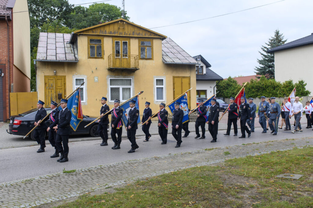 Grupa osób w mundurach niesie flagi podczas marszu ulicą. W tle żółty budynek i samochód. Dzień jest pochmurny.