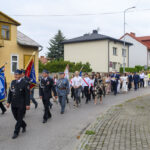 Procesja z uczestnikami idącymi ulicą w eleganckich strojach, niektórzy niosą flagi. Budynki mieszkalne w tle. Pogoda pochmurna.