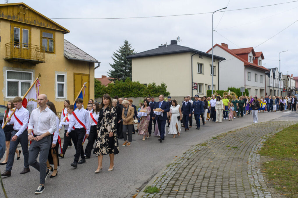 Grupa ludzi w eleganckich strojach maszeruje ulicą. Niektórzy niosą flagi, a w tle widoczne są domy. Pogoda jest pochmurna.