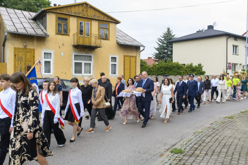 Grupa ludzi w eleganckich strojach idzie ulicą obok żółtego budynku. W tle zieleń i kolejny dom. Osoby niosą chleb na tacach.